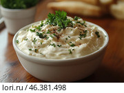 A bowl of thick, creamy sauce or dip garnished with fresh herbs such as parsley and dill and seasoned with ground black pepper. The sauce is airy and delicious, with a soft, lumpy texture. Blurred in the background are fresh herbs in a pot and breadsticks or tortillas that can be used for dipping. The light wooden table emphasizes the natural, cozy feel of the dish. Стоковое AI-изображение, AI-художник Анна Кривицкая / Фотобанк Лори