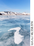 Winter landscape of frozen Baikal lake on cold sunny day in February. Snow-capped hills frame iced bay, and snow crusts in form of frozen waves form from wind on blue ice surface in mouth of Anga River. Стоковое фото, фотограф Виктория Катьянова / Фотобанк Лори