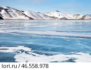 Scenic winter landscape of frozen Baikal lake on cold sunny day in February. Snow-capped hills frame iced bay, snow crusts in form of frozen waves form from wind on blue ice surface in mouth of Anga River. Стоковое фото, фотограф Виктория Катьянова / Фотобанк Лори