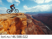 Spirit of adventure and extreme sports. A cyclist in a protective helmet and sports gear stands on the edge of a steep cliff, preparing to descend. Below, a majestic canyon with a meandering river stretches out, and snow-capped mountain peaks can be seen in the distance. The reddish rocks and deep shadows create a contrast with the bright blue sky, dotted with light clouds. The atmosphere of the photo is filled with adrenaline, freedom and a sense of endless expanses. Стоковое AI-изображение, AI-художник Анна Кривицкая / Фотобанк Лори