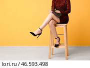 Стильная молодая женщина в блестящем красном платье и туфлях на высоком каблуке с клатчем, сидящая на барном стуле возле желтой стены. Стоковое фото, фотограф Pixel_shot / Фотобанк Лори