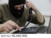 Мошенник в черной балаклаве у ноутбука с телефонной трубкой в руке. Стоковое фото, фотограф Сергей Булкин/NEWS.ru / ТАСС / Фотобанк Лори
