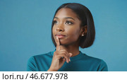 Thoughtful dark skinned woman has idea in mind, keeps finger on lips, looks pensively above, tries to decide how to act, poses against blue background. Стоковое AI-видео, аниматор Ирина / Фотобанк Лори