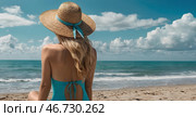 A woman on vacation, sitting in a swimsuit and a wide-brimmed straw hat on the beach on a sunny day and looking at the sea. Стоковое AI-изображение, AI-художник Photojuli / Фотобанк Лори