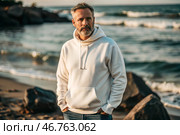 A handsome middle-aged man with a beard wearing a white hoodie on the beach. Template layout for hoodie design, print area layout for logo or design. Стоковое AI-изображение, AI-художник Photojuli / Фотобанк Лори