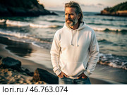A handsome middle-aged man with a beard wearing a white hoodie on the beach. Template layout for hoodie design, print area layout for logo or design. Стоковое AI-изображение, AI-художник Photojuli / Фотобанк Лори