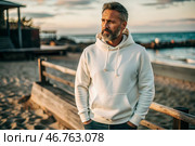 A handsome middle-aged man with a beard wearing a white hoodie on the beach. Template layout for hoodie design, print area layout for logo or design. Стоковое AI-изображение, AI-художник Photojuli / Фотобанк Лори