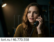 Молодая женщина внимательно разговаривает по стационарному телефону в офисе, возможно, жертва обмана. Стоковое AI-изображение, AI-художник Сергей Никифоров-Гордеев / Фотобанк Лори