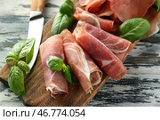 Деревянная доска с вкусными ломтиками прошутто и базиликом на столе. Стоковое фото, фотограф Pixel_shot / Фотобанк Лори