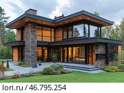 A modern country house in the style of high-tech and minimalism, surrounded by a green landscape. The house is made of glass, stone, wood and metal, creating a harmonious combination of natural and modern materials. Large panoramic windows provide an abundance of natural light and visual openness of the space. Evening lighting emphasizes the architectural lines and creates a cozy atmosphere. In the yard there is a neat lawn, ornamental plants and paved paths leading to the main entrance. Стоковое AI-изображение, AI-художник Анна Кривицкая / Фотобанк Лори