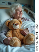 An elderly woman rests peacefully in a hospital bed, embracing a teddy bear. This moment captures the warmth of connection amidst the struggles of health challenges. Стоковое AI-изображение, AI-художник Юлия Петровская / Фотобанк Лори