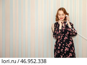A woman stands indoors, smiling as she chats on the phone. She wears a floral dress, and the pastel striped wall adds a soft backdrop to her cheerful demeanor. Стоковое фото, фотограф Aleksandr Golubev / Фотобанк Лори