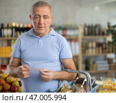 Elderly man with paper list in hands, walks around minimarket, puts groceries in market basket. Стоковое фото, фотограф Яков Филимонов / Фотобанк Лори