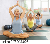 Elderly woman in company of hatha yoga lovers performs half-lotus pose, Ardha Padmasana. Стоковое фото, фотограф Яков Филимонов / Фотобанк Лори