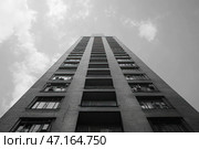 The black and white photograph captures a high-rise building, shot from the bottom up, creating an effect of perspective and infinity. The building's façade is made of brick, with rows of windows and balconies, some of which are equipped with air conditioners or grilles. The dim sky with blurred clouds gives the scene an atmosphere of urban severity and even a little melancholy. The contrast between the building and the sky emphasizes the massiveness of the structure and its impact on the surrounding space. Стоковое AI-изображение, AI-художник Анна Кривицкая / Фотобанк Лори