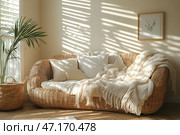 A cozy, light interior in the style of minimalism and boho. In the center of the composition is a wicker rattan sofa with soft pillows and a large knitted blanket. Warm sunlight penetrates through the blinds, creating beautiful shadows on the wall. On the floor lies a natural wicker rug, and next to the sofa is a rattan basket with a green plant. On the wall hangs a minimalist painting depicting a branch of a green plant in a wooden frame. The space radiates warmth, relaxation and harmony with nature. Стоковое AI-изображение, AI-художник Анна Кривицкая / Фотобанк Лори