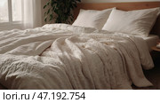 The interior of a modern house. A minimalistic cozy beige bedroom interior with a plaid, pillows, a headboard, linen linens and a bedside table. Стоковое AI-изображение, AI-художник Photojuli / Фотобанк Лори