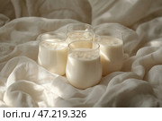 Refreshing dairy beverages served in clear glasses on soft fabric during a calm afternoon. Четыре стакана, наполненные кремообразным напитком, мирно покоятся на текстурированной мягкой ткани. Мягкий свет создает безмятежную атмосферу, идеальную для того, чтобы насладиться моментом расслабления. Стоковое AI-изображение, AI-художник Katsiaryna Repich / Фотобанк Лори
