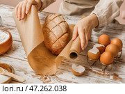 Женщина за столом заворачивает свежий хлеб в бумагу для выпечки, крупным планом. Стоковое фото, фотограф Pixel_shot / Фотобанк Лори
