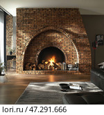 A cozy living room interior with a massive brick fireplace in the center. The fire burns in the firebox, creating a warm and relaxing atmosphere. Firewood is stacked below, with books and metal utensils nearby. The room is decorated in a loft style with elements of industrial design: brickwork, wooden floors, dark leather sofas and minimalist decor. Light pours through large windows, emphasizing the warm shade of the brick and creating a play of shadows. Стоковое AI-изображение, AI-художник Анна Кривицкая / Фотобанк Лори