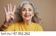 A graceful elderly woman confidently looks at the camera, resting her hands, dressed in a casual yellow t-shirt, posing indoors. Стоковое AI-видео, аниматор Ирина Старикова / Фотобанк Лори