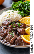 Delicious Brazilian feijoada featuring black bean stew with pork, complemented by rice, orange slices, and collard greens in festive decor. Стоковое AI-изображение, AI-художник 123 12 / Фотобанк Лори