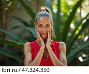 A woman showcases her confidence by touching her face and smiling, surrounded by vibrant greenery in a serene garden during daylight hours. Стоковое AI-изображение, AI-художник Sergey Grishin / Фотобанк Лори