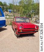 Compact red car with a distinctive unusual design. This is the SMZ S-3A — a Soviet microcar for the disabled, produced at the Serpukhov Motorcycle Plant (SMZ). The car has two seats, a very simple design and a minimalist interior. Its purpose was to provide a means of transportation for people with disabilities, especially war veterans. The car has a square shape, small wheels, headlights and a narrow front bumper. It is parked in an open area, next to other retro cars — it is probably part of an exhibition or museum space. Стоковое фото, фотограф Анна Кривицкая / Фотобанк Лори