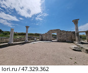 Ancient ruins on the territory of Chersonesos Taurica in Crimea. In the foreground is a rectangular square with preserved marble columns and an arch-gate in the center. These architectural elements are reminiscent of the greatness of the ancient civilization. Beyond the ruins stretches a green area and a blue sky with light clouds. All together creates an atmosphere of peace, light and deep historical significance. (2021 год). Стоковое фото, фотограф Анна Кривицкая / Фотобанк Лори