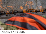 Fragment of the memorial composition: in the foreground is the St. George ribbon with its characteristic black and orange stripes, lying on flowers and fir branches. In the background is the eternal flame, a symbol of memory, respect and sorrow. The fire burns with living tongues of flame, reflected in the metal and stone of the memorial. The entire image is permeated with a solemn, solemn atmosphere, recalling the feat, sacrifice and the need to preserve historical memory for the sake of the future. Стоковое фото, фотограф Анна Кривицкая / Фотобанк Лори