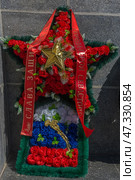 A funeral wreath in the form of a red five-pointed star, decorated with red carnations and a gold star in the center. Below, in the rectangular part of the composition, is the tricolor - the flag of the Russian Federation (white, blue, red), complemented by a decorative gold branch. The wreath is decorated with a green spruce ribbon and red mourning ribbons with the inscriptions:"Glory to the defenders" and "Hero City of Sevastopol".The composition speaks of deep respect for the fallen, the memory of heroism, patriotism and symbolizes the connection between generations. The wreath is installed near the memorial wall made of gray granite, which emphasizes the solemnity of the moment. Стоковое фото, фотограф Анна Кривицкая / Фотобанк Лори
