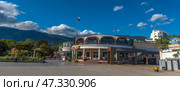 A fragment of the Yalta embankment, one of the most popular resort towns in Crimea. In the foreground is a modern building with a round façade and stained-glass windows, where cafes and restaurants are located. On the left is part of the pedestrian zone with passers-by, vacationers and strollers along the shore. In the background, the green mountains of the Crimean ridge rise, and the sky is decorated with living clouds and a flying bird. The Russian flag on the mast emphasizes the formality and maritime character of the place.The decor conveys an atmosphere of relaxation, resort activity and harmony with the natural landscape. (2021 год). Редакционное фото, фотограф Анна Кривицкая / Фотобанк Лори