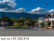 One of the central sections of the Yalta embankment overlooking a cozy park, cafes and the surrounding mountains. On the left is a well-kept green area with decorative gazebos, flowering trees and benches where people rest. In the foreground is a spacious open square with pedestrians. On the right is a modern building with a cafe and a round facade. The Russian flag flutters above it.In the distance are the densely green mountains of the southern coast of Crimea. Thick clouds hang over the peaks, creating a beautiful light and shadow effect. The atmosphere is warm, calm, resort-like. (2021 год). Редакционное фото, фотограф Анна Кривицкая / Фотобанк Лори