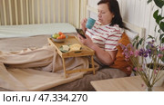 Senior woman having breakfast sitting in bed, drinking tea, enjoying her morning at home. Old lady eating from tray and using phone. Elderly female in pajamas relaxing with cup of coffee in a bedroom. Стоковое видео, видеограф Марина Демидюк / Фотобанк Лори