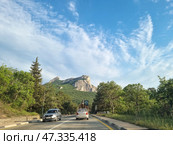 A picturesque section of the highway on the southern coast of Crimea. Ahead, a majestic view of the Kush-Kaya rock massif rises above the green forest. Cars and construction equipment move along the road. On both sides of the highway there is dense vegetation, trees, and tall grass. The sky is clear, with light cirrus clouds emphasizing the scale of the mountains and the vastness of the southern landscape. The scene conveys a sense of freedom, nature, and travel along Crimean routes. (2021 год). Редакционное фото, фотограф Анна Кривицкая / Фотобанк Лори