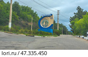Entrance sign of the resort complex "Ai-Danil", located in Crimea. The sign is made in a modern style: a large decorative structure in the form of a ring, made in beige and blue tones, inside which is the silhouette of a sailboat - a symbol of sea recreation and travel. The inscription reads: "Ai-Danil - a health resort complex". The sign is installed against the background of a green wooded slope, along the road going towards the mountains. In the frame - a cloudy sky and power lines, emphasizing the remoteness and naturalness of the area. Стоковое фото, фотограф Анна Кривицкая / Фотобанк Лори