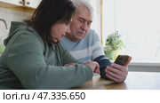 Old couple using a phone together while relaxing in the kitchen at home. Elderly man and woman spending leisure time browsing online with a smartphone. Happy senior married couple typing, learning. Стоковое видео, видеограф Марина Демидюк / Фотобанк Лори