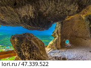 One of the cave halls of the Chelter-Marmara Monastery in Crimea. The view extends through natural arches and niches in the rock - as if nature itself carved this ancient temple. From the inside, a panoramic view of green hills, forests and distant mountain ranges opens up. Light streams in through a round hole in the wall, creating a play of shadows and emphasizing the relief of the rock surfaces, smooth from time and wind.The cave seems to breathe history - in its silence, the presence of centuries is felt. This is not just a shelter, but a place of spiritual solitude, prayer and observation of the world from above. Natural architecture combines with the harsh beauty of the Crimean landscapes, creating a unique atmosphere of sacred solitude. Стоковое фото, фотограф Анна Кривицкая / Фотобанк Лори