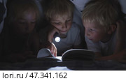 Three young children are reading a book with a flashlight under the covers at night. Стоковое видео, видеограф Mikhail Davidovich / Фотобанк Лори