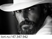Expressive black and white portrait of a man in a wide-brimmed light-colored hat. His face is covered with light stubble and a thick beard, and his gaze is piercing, confident, almost hypnotic. The deep texture of the skin and hair is emphasized by the play of light and shadow, creating a cinematic effect. The composition is built very close to the face, which increases the tension and charisma of the image. The image is associated with a western, Latin American aesthetics or classic noir style. Стоковое AI-изображение, AI-художник Анна Кривицкая / Фотобанк Лори