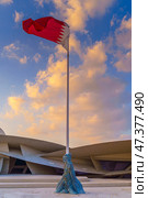 Doha, Qatar - Jan 06 2024, a view of flagpole with the flag of Qatar, fluttering in the wind, with the National Museum of Qatar on background, at evening with cloudy sky, Doha, Qatar. Редакционное фото, фотограф Сергей Богомяко / Фотобанк Лори