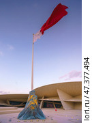 Doha, Qatar - Jan 06 2024, a view of flagpole with the flag of Qatar, fluttering in the wind, with the National Museum of Qatar on background, at evening with cloudy sky, Doha, Qatar. Редакционное фото, фотограф Сергей Богомяко / Фотобанк Лори