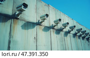 Surveillance cameras on a concrete wall, symbolizing border control and security. Ряды камер наблюдения, установленных на высокой бетонной стене, символизирующей пограничный контроль и безопасность. Стоковое AI-изображение, AI-художник Katsiaryna Repich / Фотобанк Лори