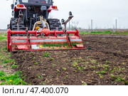 A tractor equipped with a red rotary cultivator actively prepares the soil in a rural field. The equipment loosens the earth, preparing it for planting and cultivation. Стоковое фото, фотограф Коханова Наталья / Фотобанк Лори