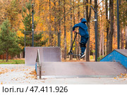 A teenager on a stunt scooter on a playground in a skate park. A child in a tracksuit performs a trick in an autumn park while jumping. Стоковое фото, фотограф Photojuli / Фотобанк Лори