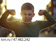 Kid boy shows his muscles. Angry little child boy waves his fist at the camera outdoors at the city street. Unhappy grumpy boy showing his biceps. Provocation. Elementary school problems. Bullying. Стоковое AI-изображение, AI-художник Марина Демидюк / Фотобанк Лори