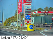 Lukoil gas station on the side of the road. In the foreground are road signs with directions, a bright yellow bollard with a blue pointer and a digital board with fuel prices (DT, 92, 95, 100, EKTO). In the background are gas stations, a truck with a tarpaulin body and several cars. Red Lukoil flags and a building with a minimarket and a cafe are visible. Autumn foliage of the trees adds warmth and depth to the composition, and the clear blue sky creates a bright contrast. (2020 год). Редакционное фото, фотограф Анна Кривицкая / Фотобанк Лори