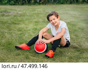 Улыбающийся мальчик-футболист с красным футбольным мячом, в красных ярких бутсах в футбольной форме и носках сидит на зеленой траве. Стоковое фото, фотограф Елена Болтунова (Галиуллина) / Фотобанк Лори