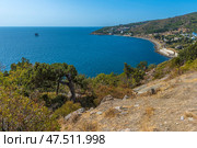 A magnificent view of the Crimean coast. From above, a panorama of the curved coastline, covered with sparse vegetation, rocks and junipers, opens up. In the distance, a small town or village with white houses spread out along the slope of a hill is visible. The Black Sea is deep blue, calm, with slight glare from the sun. On the horizon, closer to the center of the frame, an old sea platform or pier is noticeable. This place conveys an atmosphere of privacy, tranquility and southern warmth. Стоковое фото, фотограф Анна Кривицкая / Фотобанк Лори