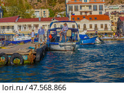 A picturesque scene on the Balaklava embankment: fishermen and locals at the pier prepare boats tied to a wooden pontoon. In the frame - traditional motor boats, the architecture of a southern city and clean sea water. The atmosphere of coziness, resort life and authentic sea life perfectly conveys the spirit of the south and the measured rhythm of a seaside village. (2020 год). Редакционное фото, фотограф Анна Кривицкая / Фотобанк Лори